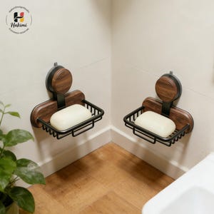 Pode incluir: Duas saboneteiras de parede. Cada uma apresenta uma base de madeira escura e uma cesta de arame de metal preto. Um detalhe redondo de madeira escura está acima da cesta. Cada uma contém uma barra de sabão branca. A marca "Hakimi" está no canto superior esquerdo.
