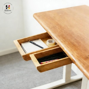 May include: A light brown wooden desk with two pull-out drawers. The top drawer contains a notepad, pen, and tape roll. The bottom drawer holds pencils. The desk has a white base, suitable for a home office or study.