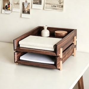 K&ouml;nnte beinhalten: Ein zweistufiger Schreibtisch-Organizer aus Holz mit dunkelbraunem Finish. Der Organizer enth&auml;lt einen Stapel wei&szlig;es Papier und einen Stapel wei&szlig;er B&uuml;cher. Oben stehen eine kleine wei&szlig;e Vase und eine Holzkiste. Der Organizer steht auf einer wei&szlig;en Oberfl&auml;che.