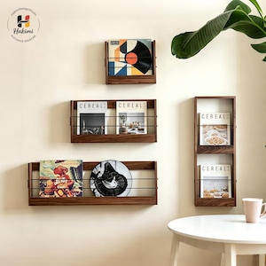 May include: Four dark wood wall-mounted display shelves with thin black metal bars across the front. The shelves hold various vinyl records and Cereal magazines. The top shelf holds one record, the middle holds two magazines, the bottom holds a record and a circular disc, and the vertical shelf holds two magazines.