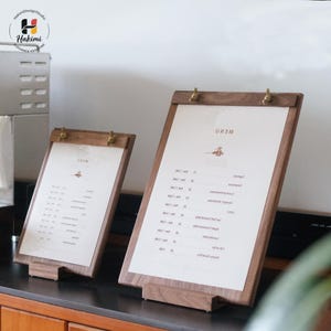 May include: Two wooden menu holders with brass clips and a stand. Each holder displays a printed menu with text and a small illustration. The wood has a warm, natural tone, and the holders are angled for easy viewing.