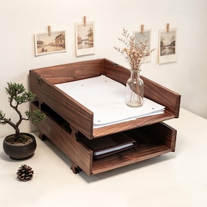 May include: A two-tiered wooden desk organizer with a dark brown finish. The top tray holds white paper, and the bottom tray contains a brown notebook. A small bonsai tree and a glass vase with dried flowers are also present.