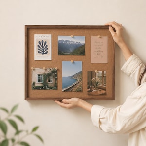 May include: A rectangular cork bulletin board with a dark wood frame. Five items are pinned to the board: a Matisse print, a mountain landscape photo, a card with text, a photo of a house with vines, a coastal train track photo, and a floral still life.