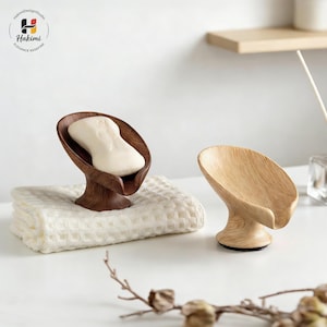 May include: Two wooden soap dishes. One is dark brown with a bar of soap, the other is light brown. Both have a leaf-like design and a pedestal base. The dark brown soap dish sits on a white waffle-weave towel.