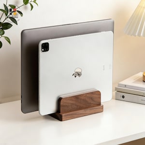 May include: A wooden tablet and laptop stand in a natural wood finish. The stand holds a silver tablet and a gray laptop. The stand is on a white surface. The Apple logo is visible on the tablet.