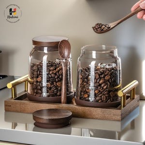 May include: Two glass coffee bean jars with wooden lids and measuring lines sit on a wooden tray with brass handles. A wooden spoon holds coffee beans. The jars are filled with coffee beans.