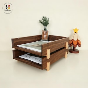 K&ouml;nnte beinhalten: Ein zweistufiger, dunkelbrauner Schreibtisch-Organizer aus Holz mit hellen Holzst&uuml;tzen. Das obere Fach enth&auml;lt eine Zeitschrift. Eine kleine Glasvase mit Gr&uuml;n und ein dekorativer Baum aus Holz sind ebenfalls vorhanden.
