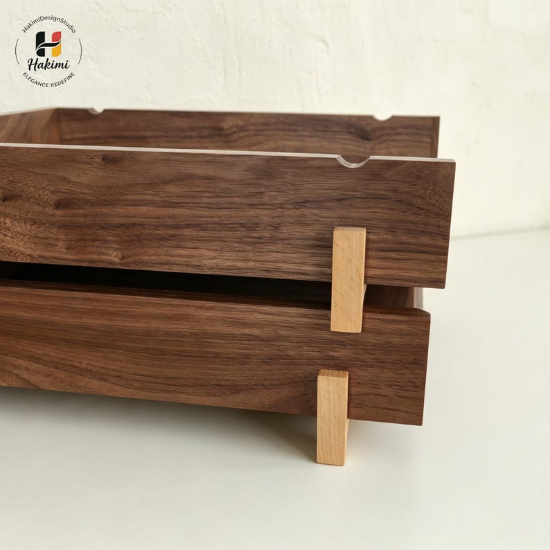 K&ouml;nnte beinhalten: Zweistufiges Holztablett mit dunkelbraunem Finish. Das Tablett besteht aus dicken Holzbrettern und verf&uuml;gt &uuml;ber hellfarbene Holzst&uuml;tzen. Das obere Tablett hat einen kleinen Ausschnitt am Rand. Das Logo "Hakimi Design Studio" befindet sich in der oberen linken Ecke.