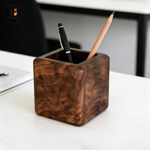 May include: A square, wooden desk organizer with a rich, dark brown grain pattern. It holds a black pen and a wooden pencil. The organizer sits on a white surface, suggesting a workspace or office setting.