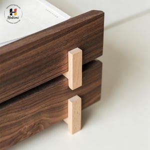 K&ouml;nnte beinhalten: Nahaufnahme eines zweistufigen Schreibtisch-Organizers aus Holz. Der Organizer besteht aus dunkelbraunem Holz mit hellen Holzst&uuml;tzen. Auf der oberen Ebene liegt ein Stapel wei&szlig;es Papier. Das Logo "Hakimi Design Studio" befindet sich in der oberen linken Ecke.