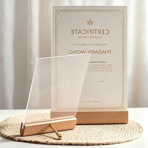 May include: A clear acrylic sign holder with a light-toned wooden base and a gold-colored metal support pin. Behind it stands a vertical certificate of appreciation with a crown icon and the printed text Certificate of Appreciation.