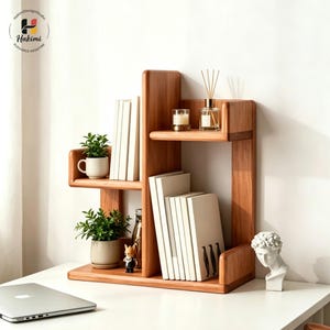 May include: A wooden desk organizer with multiple shelves, holding books, small potted plants, and decorative items. The organizer is made of light-colored wood and has a modern design. A laptop is visible on the desk. The brand name "Hakimi" is visible in the top left corner.