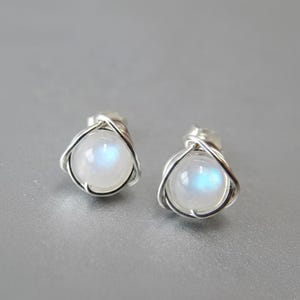 May include: A pair of silver stud earrings featuring a round, iridescent moonstone cabochon. The moonstone is cradled within a twisted silver wire setting, creating a delicate and elegant design. The earrings have a subtle, ethereal glow.