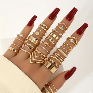 May include: A collection of gold-colored rings in various designs, including bands, geometric shapes, and rings with small gemstones. The rings are displayed on fingers with long, red nails, creating a stylish and fashionable look. The rings are a mix of textures and styles.