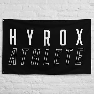 May include: A black rectangular flag featuring the white text HYROX in a bold, sans-serif font on the top line and the white outlined text ATHLETE in a matching font on the bottom line. The flag has metal grommets in the corners for hanging.