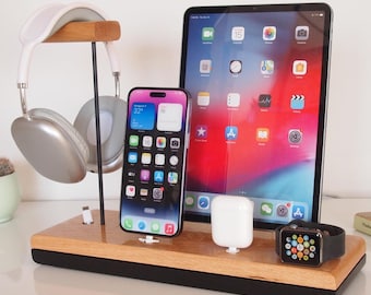 May include: A wooden charging station with a black base, holding a smartphone, headphones, a tablet, wireless earbuds, and a smartwatch. The station is on a white surface.