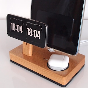Peut inclure: Station de charge en bois avec une horloge numérique affichant 18h04. Il contient une tablette et un téléphone, ainsi qu'un socle de chargement sans fil blanc pour écouteurs. La base est en bois brun clair avec un bord noir.