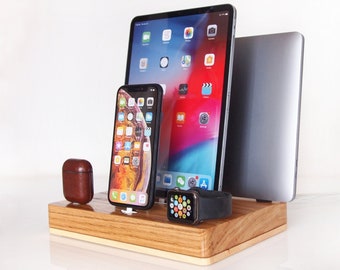 May include: A rectangular wooden docking station featuring slots to hold a tablet, a smartphone, a laptop, and a smartwatch. A brown leather case for wireless earbuds sits on the left side of the base, while the devices are arranged upright.