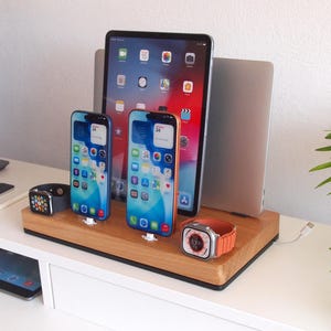 May include: A wooden charging station holds two smartphones, a tablet, and a laptop. Two smartwatches are also on the station. The phones and watches are charging. The station is on a white surface.