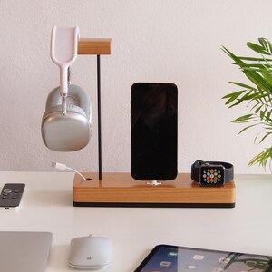 May include: A wooden charging station with a black base holds a smartphone, smartwatch, and silver headphones. A white charging cable is connected to the headphones. A small plant is in the background.