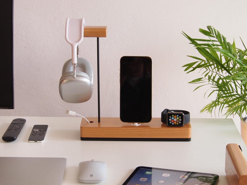 May include: A wooden charging station with a black base holds a smartphone, smartwatch, and silver headphones. A white charging cable is connected to the headphones. A small plant is in the background.