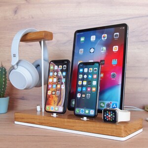 May include: A wooden desk organizer with a white base holds a tablet, two smartphones, and a smartwatch. A pair of gray headphones hangs from a wooden stand. A small cactus in a blue pot is visible on the left.