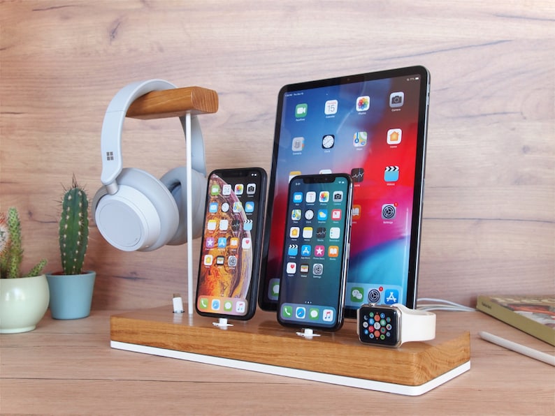 May include: A wooden desk organizer with a white base holds a tablet, two smartphones, and a smartwatch. A pair of gray headphones hangs from a wooden stand. A small cactus in a blue pot is visible on the left.
