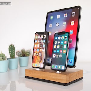 Può includere: Una stazione di ricarica in legno con tre slot per caricare telefoni e tablet. La stazione ha una base nera e un piano in legno chiaro. Ci sono tre dispositivi sulla stazione: un tablet e due telefoni. Il tablet è in modalità verticale e i telefoni sono in modalità orizzontale.
