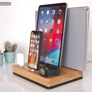 Peut inclure: Station de charge en bois avec emplacements pour tablette, smartphone et montre connectée. La station est en bois clair avec une base noire. Un ordinateur portable argenté est également en arrière-plan. L'écran de la tablette affiche diverses icônes d'applications.