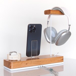 May include: Wooden charging station with a white base, holding a black smartphone, a white smartwatch, and silver headphones. The headphones hang from a wooden bar. A charging cable is visible.