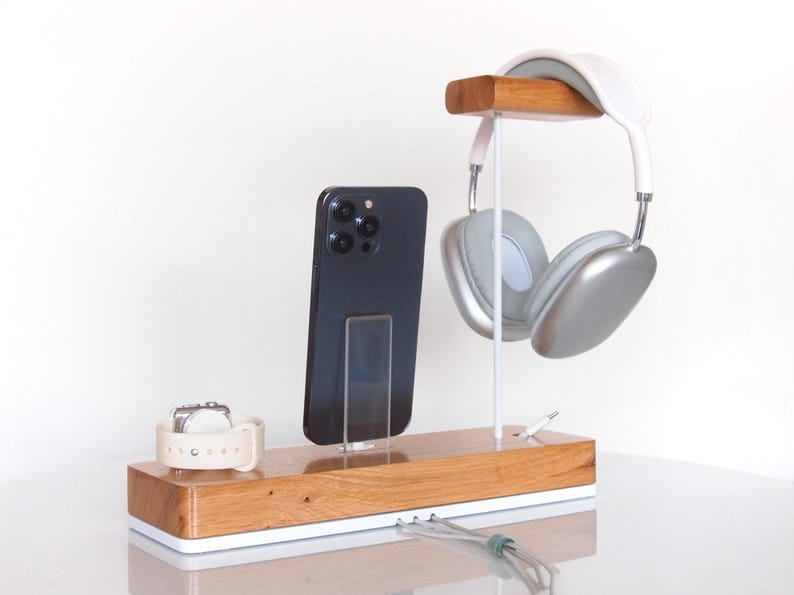 May include: Wooden charging station with a white base, holding a black smartphone, a white smartwatch, and silver headphones. The headphones hang from a wooden bar. A charging cable is visible.