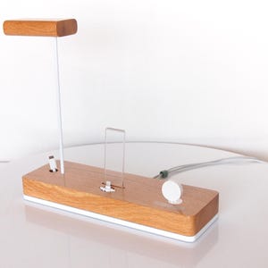 May include: A wooden charging station with a light brown finish and a white base. It features a vertical stand with a wooden top, a clear acrylic phone holder, and a white circular watch charger. A white cable extends from the station.