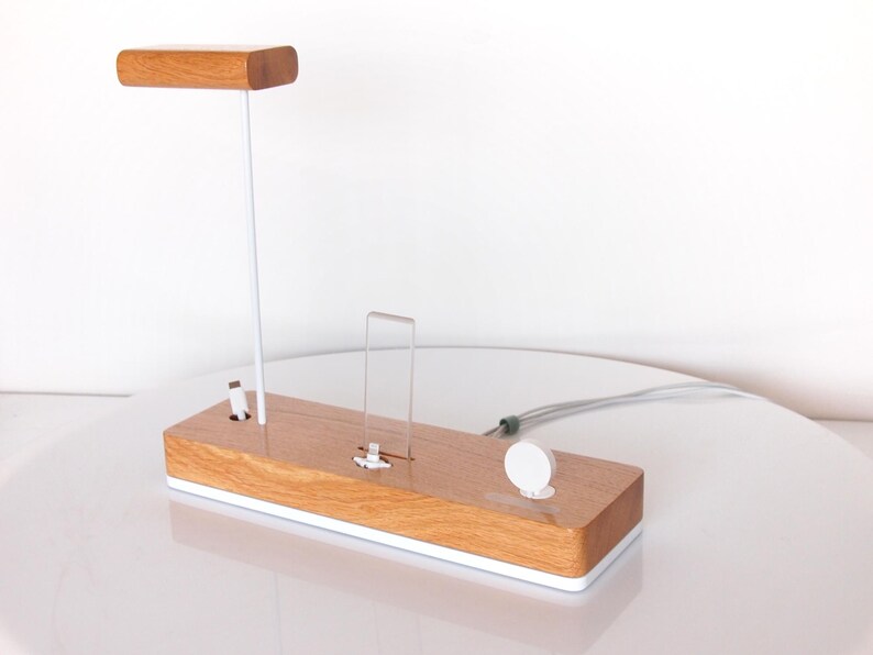 May include: A wooden charging station with a light brown finish and a white base. It features a vertical stand with a wooden top, a clear acrylic phone holder, and a white circular watch charger. A white cable extends from the station.