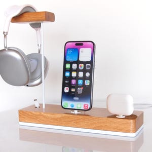 May include: A wooden desk organiser with a phone, headphones, and wireless earbuds. The phone is black with a colourful app display. The headphones are silver and white, and the earbuds are white. The organiser has a white base.