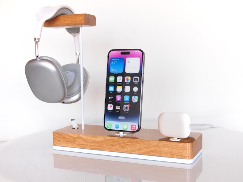 May include: A wooden desk organiser with a phone, headphones, and wireless earbuds. The phone is black with a colourful app display. The headphones are silver and white, and the earbuds are white. The organiser has a white base.