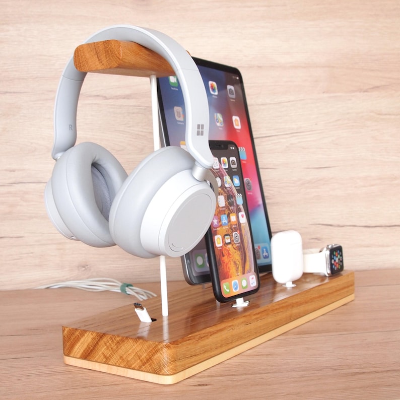 May include: A wooden charging station with a light gray headset, a tablet, a smartphone, a wireless charging pad, and a smartwatch. The station is made of light brown wood and has multiple slots for devices.