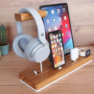 May include: A wooden desk organizer with a white trim holds a tablet, smartphone, wireless earbuds, and a smartwatch. A pair of light blue headphones rests on a wooden bar. The organizer is on a wooden desk.