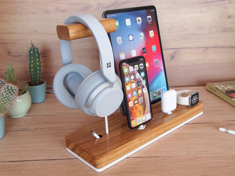 May include: A wooden desk organizer with a white trim holds a tablet, smartphone, wireless earbuds, and a smartwatch. A pair of light blue headphones rests on a wooden bar. The organizer is on a wooden desk.