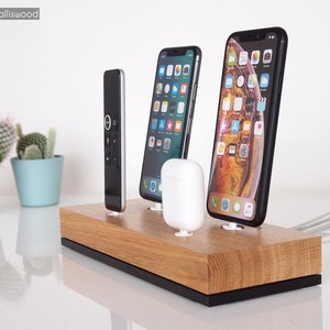 Könnte beinhalten: Eine Holz-Ladestation mit schwarzem Sockel, die zwei iPhones, eine Fernbedienung und ein Paar weiße kabellose Ohrhörer hält. Die Station steht auf einer weißen Oberfläche.