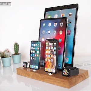 May include: A wooden charging station holds multiple electronic devices, including smartphones, tablets, and smartwatches. The station is made of light-colored wood and has slots for each device. The devices are black and silver, and the smartwatches have black bands.