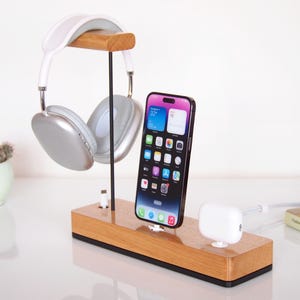 May include: Wooden headphone and phone charging station. The station holds silver and white headphones, a black smartphone, and a white charging dock. The wooden base has a rectangular shape.