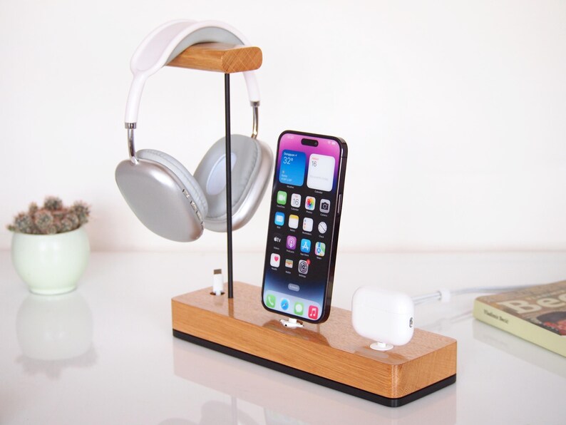 May include: Wooden headphone and phone charging station. The station holds silver and white headphones, a black smartphone, and a white charging dock. The wooden base has a rectangular shape.