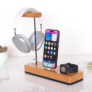 May include: A wooden charging station holding a phone, smartwatch, and headphones. The phone has a colourful display. The headphones are silver and white. The smartwatch is black with a silver case. A small succulent plant in a white pot is to the left.