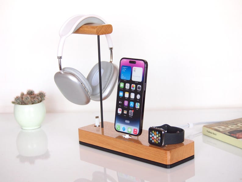 May include: A wooden charging station holding a phone, smartwatch, and headphones. The phone has a colourful display. The headphones are silver and white. The smartwatch is black with a silver case. A small succulent plant in a white pot is to the left.
