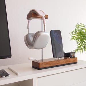 May include: A wooden charging station with a headphone stand, phone charger, and watch charger. The headphones are silver and white. The phone screen displays 03:25. A white cable is plugged into the base. A small plant in a white pot is in the background.