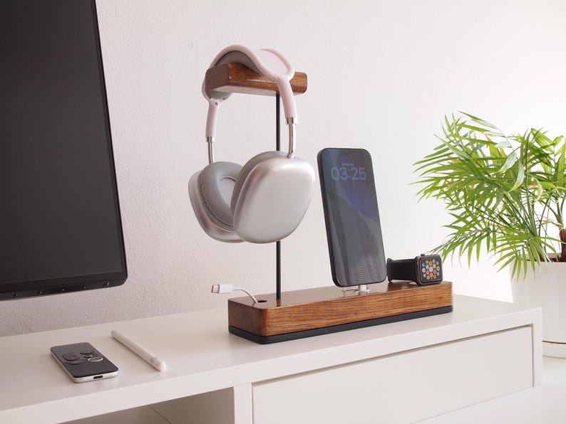 May include: A wooden charging station with a headphone stand, phone charger, and watch charger. The headphones are silver and white. The phone screen displays 03:25. A white cable is plugged into the base. A small plant in a white pot is in the background.