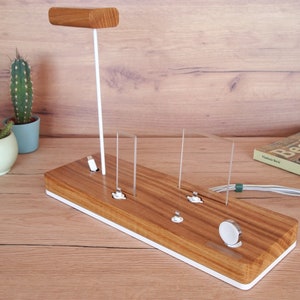 May include: Wooden charging station with a white base and accents. Features a vertical stand with a wooden top, and multiple charging ports for various devices. Includes a spot for a smartwatch. The station is on a wooden surface.