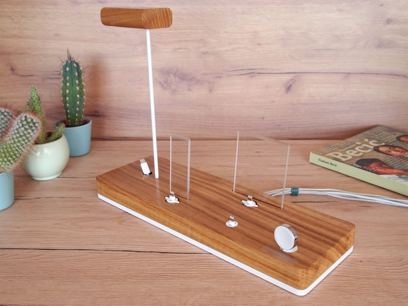 May include: Wooden charging station with a white base and accents. Features a vertical stand with a wooden top, and multiple charging ports for various devices. Includes a spot for a smartwatch. The station is on a wooden surface.
