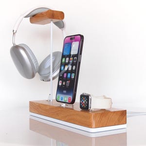 May include: Wooden charging station with a white base, holding a smartphone, smartwatch, and silver headphones. The phone is black and has a colourful display. The headphones are suspended above the phone. The smartwatch has a white band.