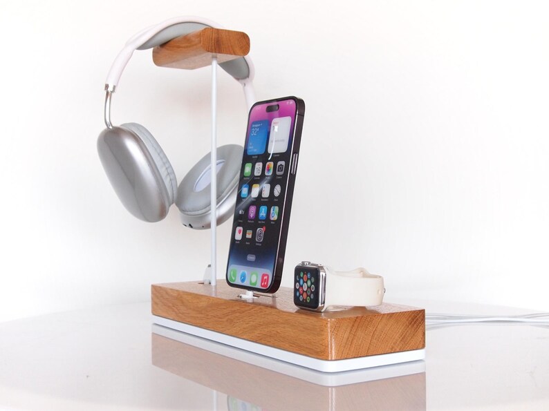 May include: Wooden charging station with a white base, holding a smartphone, smartwatch, and silver headphones. The phone is black and has a colourful display. The headphones are suspended above the phone. The smartwatch has a white band.